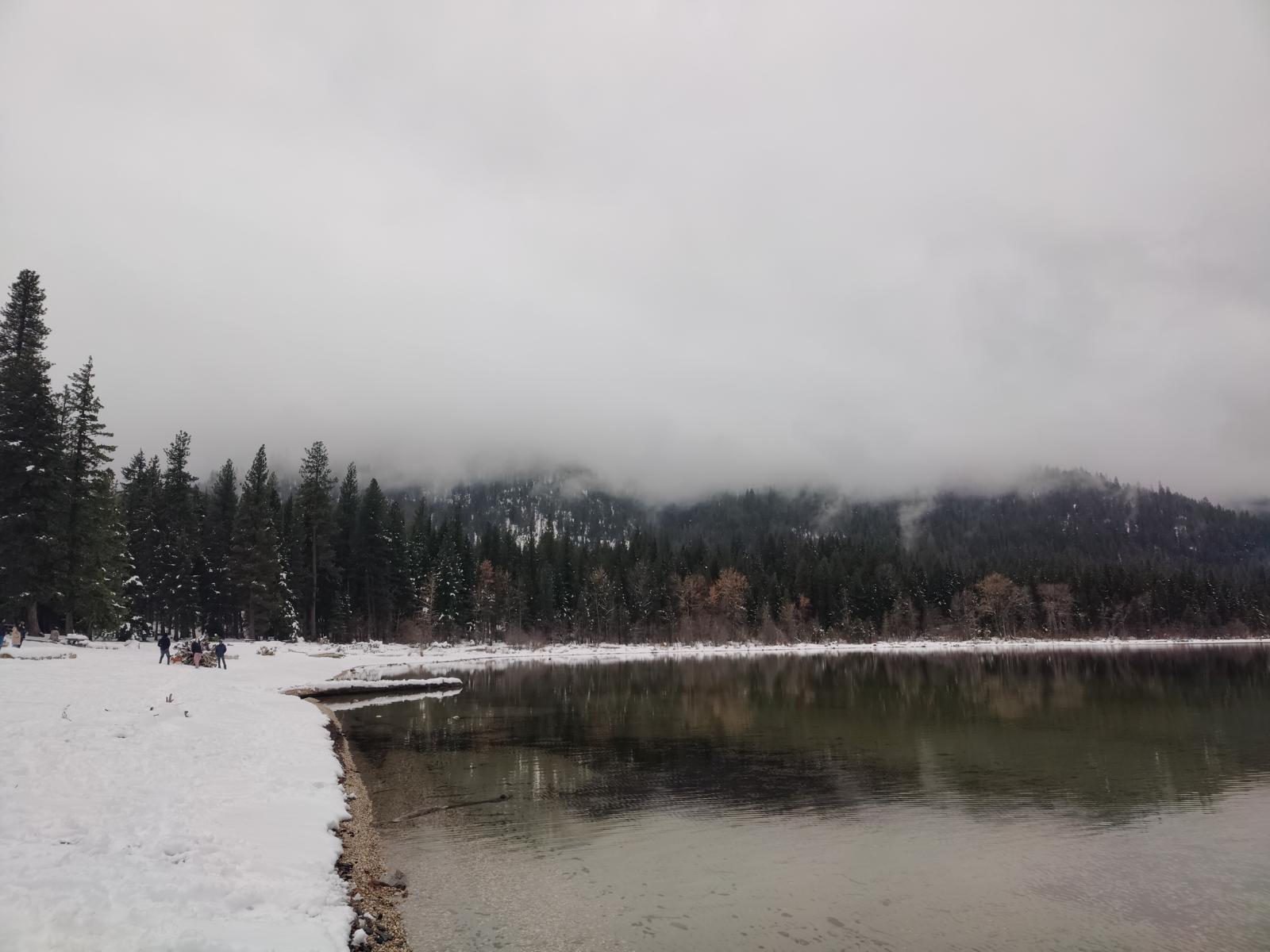 Snow-edged lake beneath foggy evergreen-covered hills